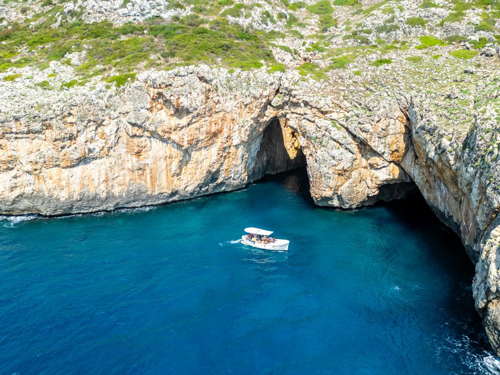 Küste Apuliens in der Nähe von Santa Maria di Leuca, ruhige Wellen und felsige Ufer; ein Boot fährt entlang der Küste; aufgenommen mit Drohne